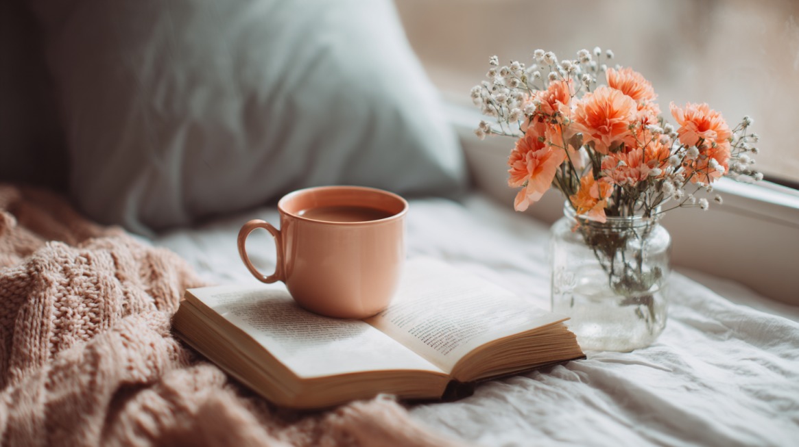 Cozy scene with a cup of coffee resting on an open book beside a vase of soft orange flowers by the window